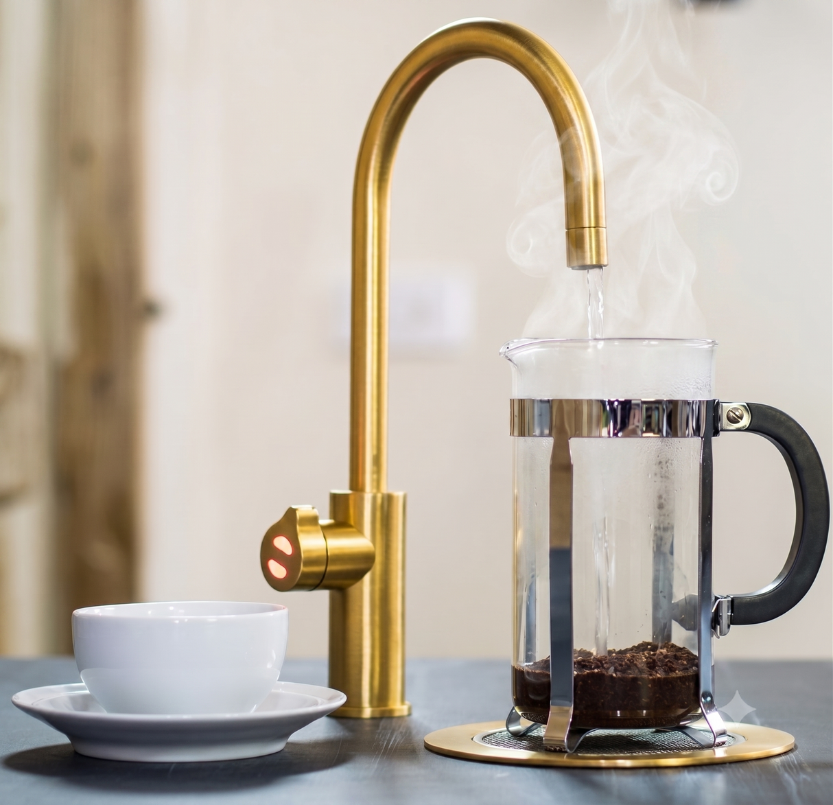 A modern curved brass faucet with a side temperature knob, shown dispensing hot water into a glass coffee maker with visible steam. A white cup and saucer sit on the counter beside it, with a softly blurred indoor background.