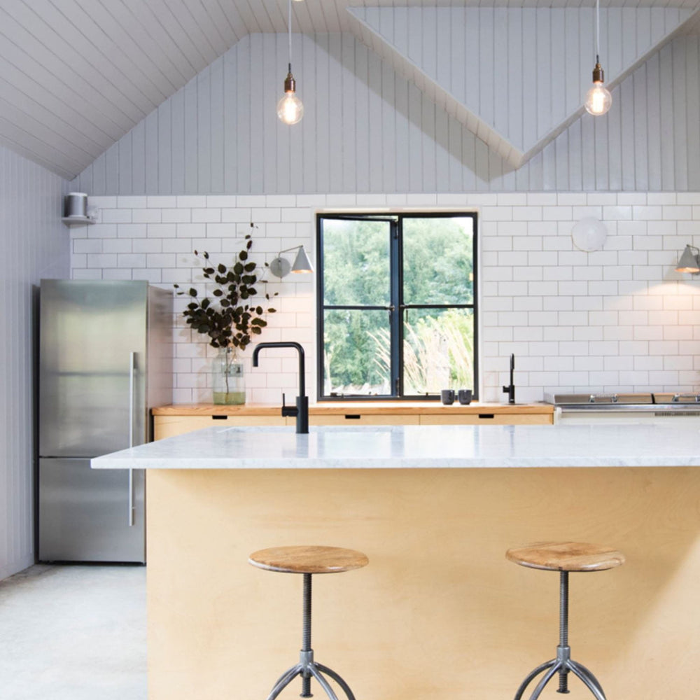 A modern kitchen interior with a light wood island, white subway tile backsplash, stainless steel refrigerator, black faucet, and two wooden-topped bar stools in front of the island.