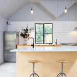 A modern kitchen interior with a light wood island, white subway tile backsplash, stainless steel refrigerator, black faucet, and two wooden-topped bar stools in front of the island.