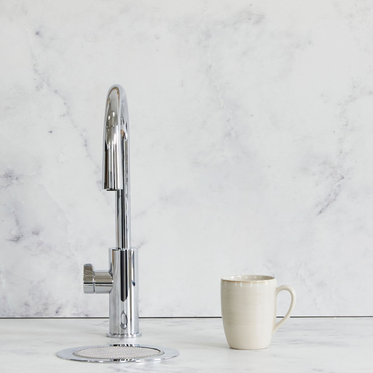 A modern chrome high-arc single-handle faucet with a circular drain base, shown next to a beige ceramic mug on a white marble countertop against a matching marble backsplash.
