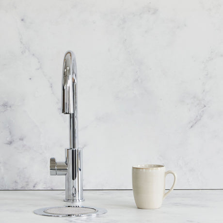 A modern chrome high-arc single-handle faucet with a circular drain base, shown next to a beige ceramic mug on a white marble countertop against a matching marble backsplash.